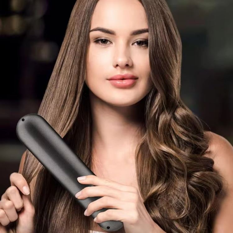 A woman with long, wavy brown hair uses the UNIQ 2-in-1 Straightening Brush with Ceramic Plates—featuring fast heating and USB-C charging—to style her hair. She looks ahead with a neutral expression; the background is blurred.