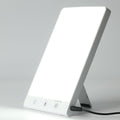Die Fashiongirl Lichttherapie-Lampe 10.000 Lux LED ist eine rechteckige Lampe mit einem hellweißen Bildschirm, einer ausklappbaren Halterung, drei Bedientasten und einem Netzkabel an der Unterseite – entwickelt, um künstliches Tageslicht zu simulieren.