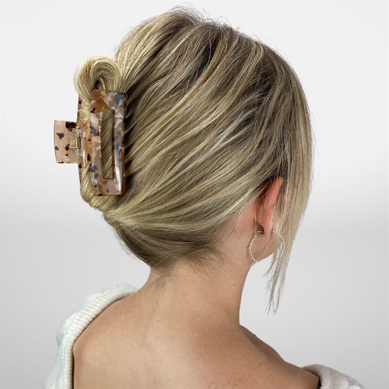 A woman with thick blonde hair styled in a low twist, secured by the SOHO Aria Hair Claw in Light Tortoise, wears silver hoop earrings and a white top, facing away from the camera against a plain background.