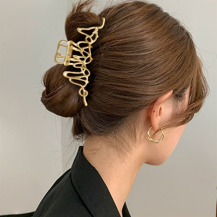 A woman with brown hair in a low bun, secured with the SOHO Asli Metal Hair Claw - Silver by SOHO. She wears a black top and gold twisted hoop earring. The photo, taken from behind and the side, highlights her elegant silver hair accessory.