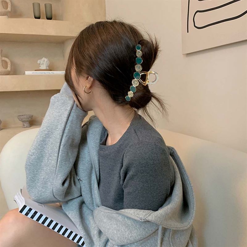 A woman with brown hair sits on a couch, facing away. She wears a grey sweater and secures her hair with the SOHO Becca Hair Claw - Lime by SOHO, featuring green and gold circles. She holds a striped notebook and rests her head on her hand.