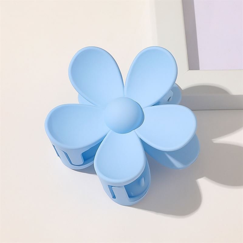 The SOHO Bloom Hair Claw in light blue, a stylish flower-shaped hair accessory by SOHO with six rounded petals, rests on a white surface beside a white picture frame—an irresistibly cute and trendy hair clip.