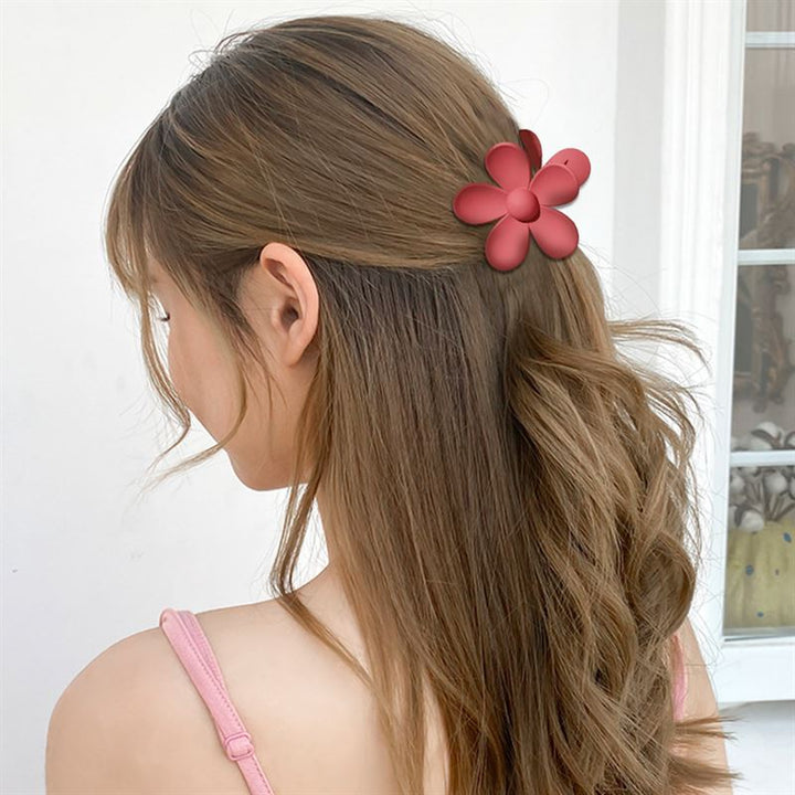 A woman with long, wavy brown hair is seen from behind wearing the SOHO Bloom Hair Claw - Pink Ombre by SOHO, paired with a light pink sleeveless top. A white wall and window are visible in the background.