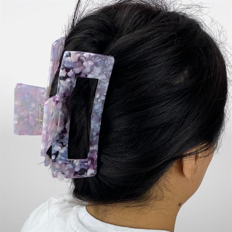 A person with dark hair wears the SOHO Clara Hair Claw - Lavender Pink by SOHO, styled to hold their hair up. They wear a white top against a plain white background.