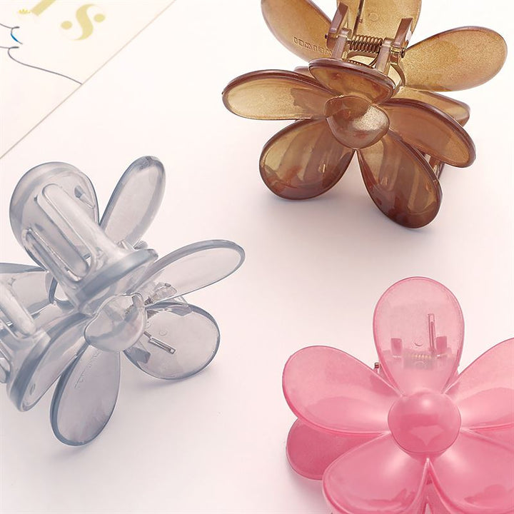 The SOHO Fiore Hair Claw - Russet by SOHO is a flower-shaped hair accessory with translucent petals and a round center, shown in brown, gray, and pink on a light surface near the edge of white paper.