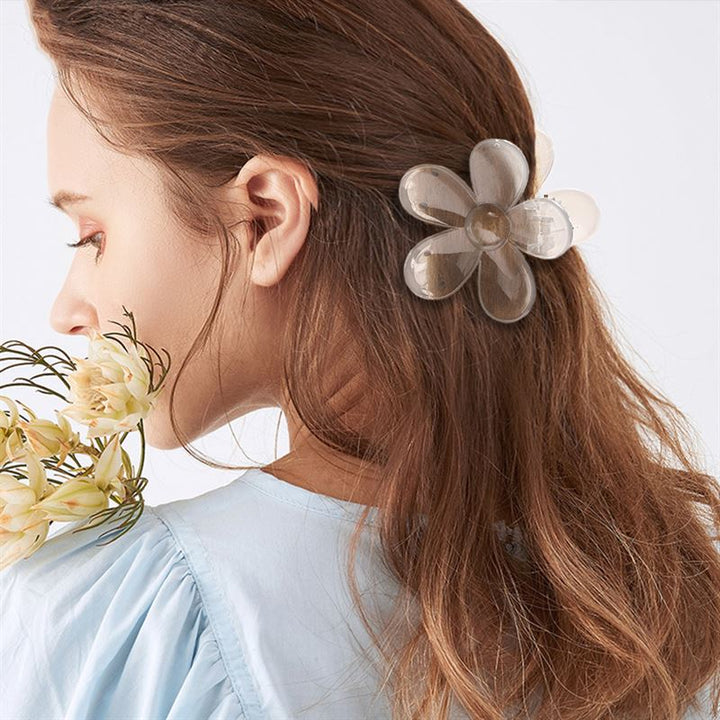 A woman with long brown hair wears the SOHO Fiore Hair Claw - Russet by SOHO and holds pale yellow flowers near her face. She is dressed in a light blue top and is shown from the side against a plain background.
