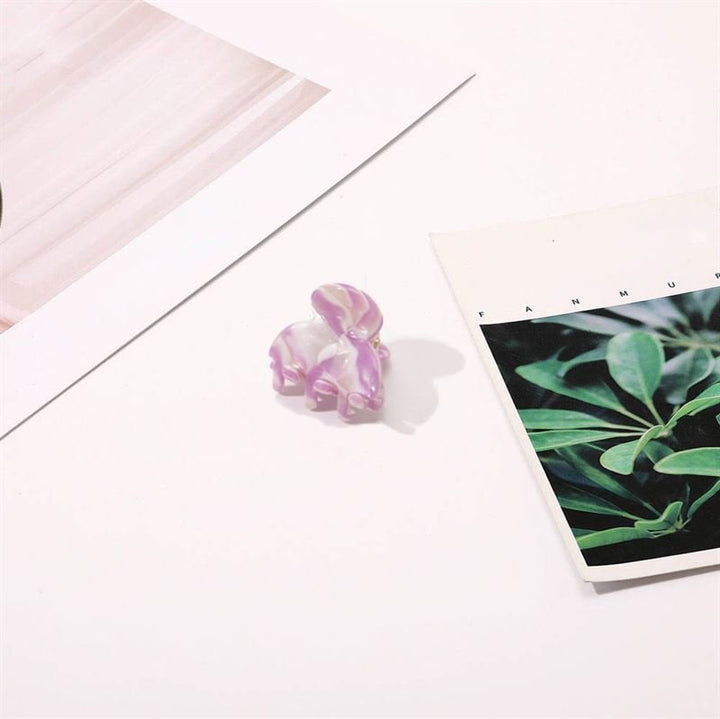 The SOHO Hara Mini Hair Claw in Periwinkle—a small, butterfly-shaped hair accessory with marbled purple and white hues—rests on a white surface near two sheets of paper: one with green leaves, the other striped.
