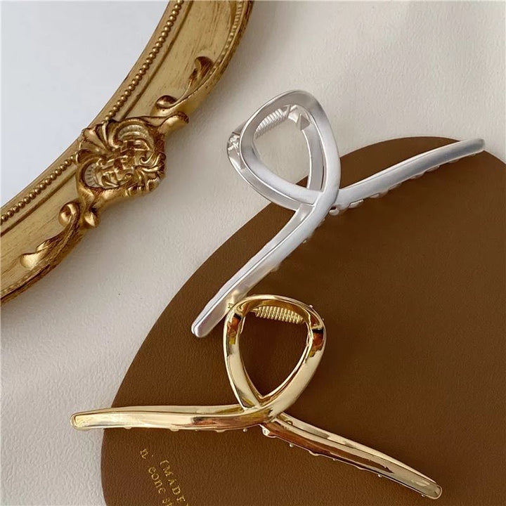 The SOHO Iva Metal Hair Claw in Silver by SOHO is displayed on a brown surface near an ornate gold-framed mirror. Its modern, twisted design is perfect to secure a messy bun or elevate any elegant hairstyle.
