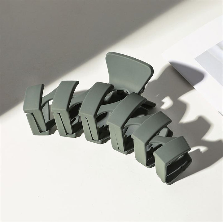 The SOHO Just Hair Claw - Green by SOHO, a large matte olive hair clip with wide rectangular teeth, sits on a white surface near partially shadowed paper showing a gray section.