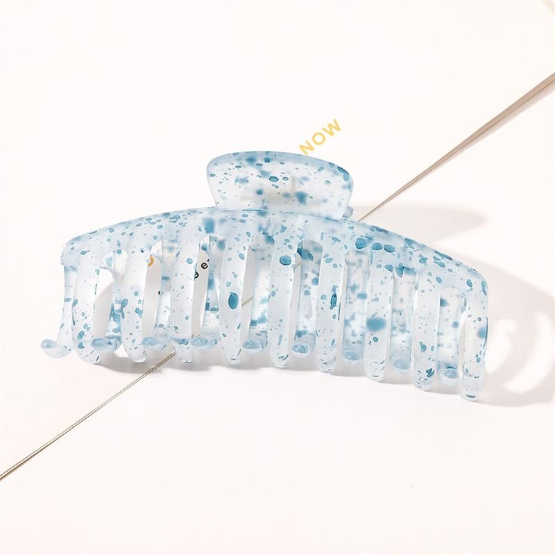 The SOHO Kana Splash Hair Claw - Blue by SOHO, featuring a translucent blue and white marbled design with a speckled pattern, offers a strong grip for all hair types. It rests on a white surface with "NOW" partially visible in the background.