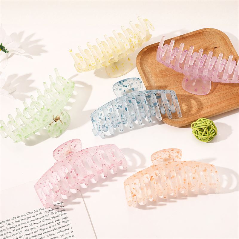 The SOHO Kana Splash Hair Claw in Purple features a pastel-speckled design and a strong grip for all hair types. Displayed on a white surface with a wooden tray, green decor ball, and open magazine nearby.