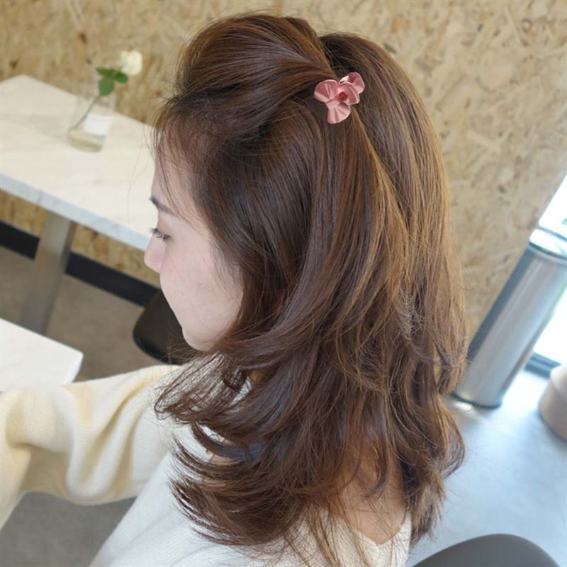 A woman with shoulder-length brown hair, wearing a cream sweater, styles her hair with the SOHO Lala Mini Hair Claws - Pastel Flowers. She sits at a table with a white table and a vase of flowers in the background.