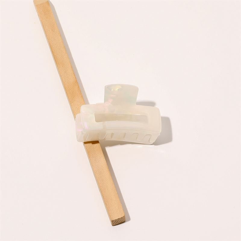 The SOHO Leia Mini Hair Claw - Pearl by SOHO, a translucent iridescent accessory, is clipped onto a slim wooden stick against a light background—perfect for adding effortless elegance to your hairstyle.