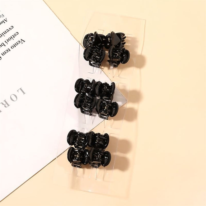 A clear plastic strip holds ten SOHO Lucca Mini Hair Claws - Black by SOHO, displayed on a light beige surface beside a partially visible white sheet with black text.