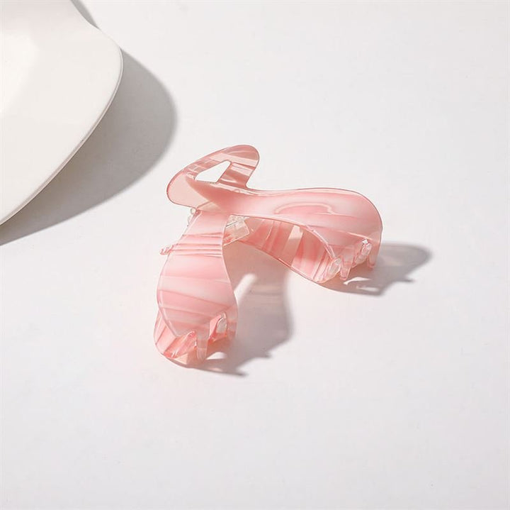 The SOHO Malou Hair Claw - Pink by SOHO, featuring a translucent pink marbled design, rests on a white surface near the edge of a white dish, casting a soft shadow.