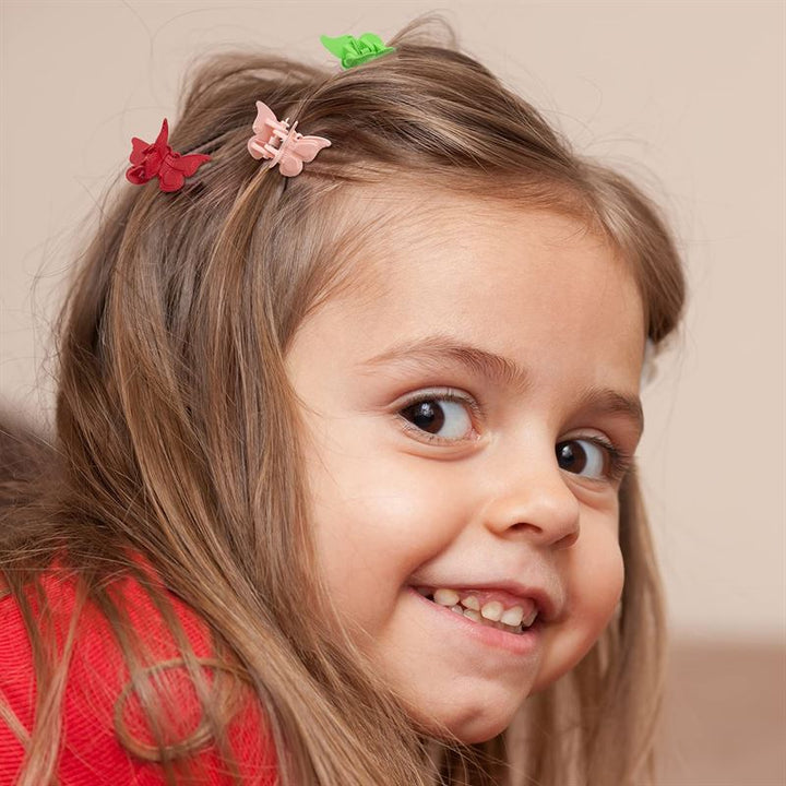 A young girl with light brown hair smiles, wearing SOHO Mini Butterfly Hair Claws—50 multicolour hair decorations by SOHO. She’s in a red top, looking slightly over her shoulder at the camera.