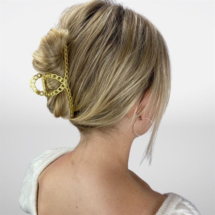 A woman with blonde hair in a low twist, secured by the SOHO Monz Metal Hair Claw in Gold from SOHO. She wears a white sweater and large hoop earrings, with her back to the camera.