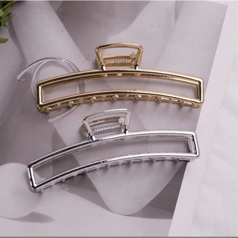The SOHO Selin Metal Hair Claw in Silver by SOHO is a minimalist, rectangular curved hair clip with an open frame and visible spring, designed to suit all hair types and effortlessly secure a messy bun. Shown on a black-and-white background.