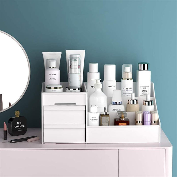 The UNIQ Cosmetic Organiser with Two Drawers, White - P110 displays skincare bottles, creams, and sprays on a pink vanity next to a round mirror and black Chanel perfume. A teal wall in the background adds vibrant color.