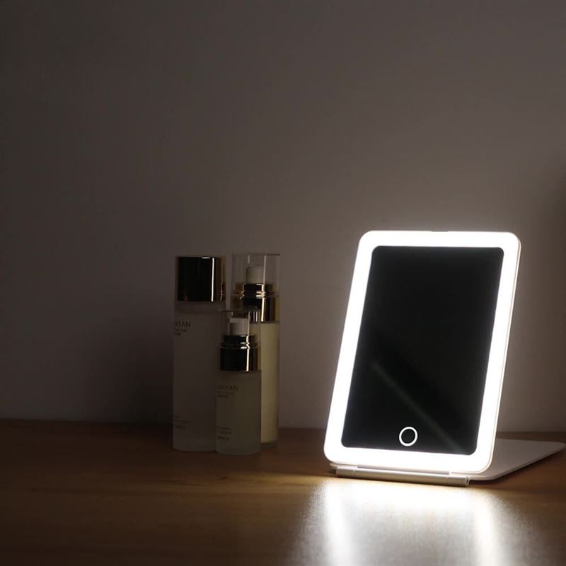 The UNIQ Foldable LED Makeup Mirror—rechargeable, travel-friendly, and white—sits illuminated on wood next to three skincare bottles against a plain wall.