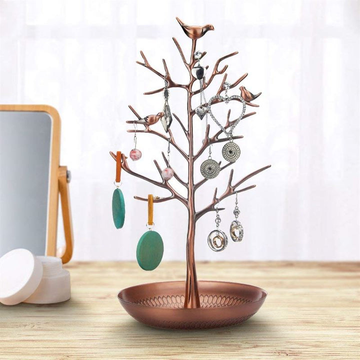 The UNIQ Bronze Vintage Jewelry Tree with 3 Birds elegantly displays earrings on its branches, serving as a stylish organizer stand beside a small mirror and round white containers on a wooden surface.